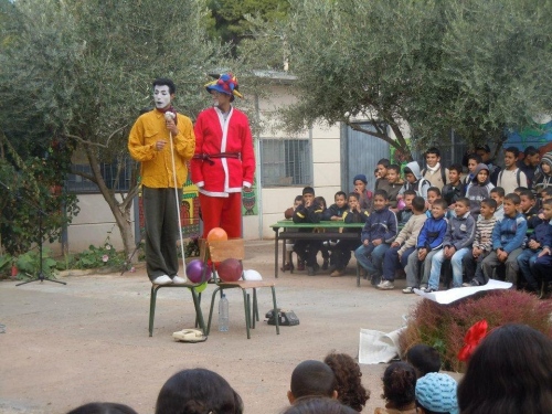 Two clowns stand on a chair in the village square, with somewhat esoteric props including old telephones and a length of rope.